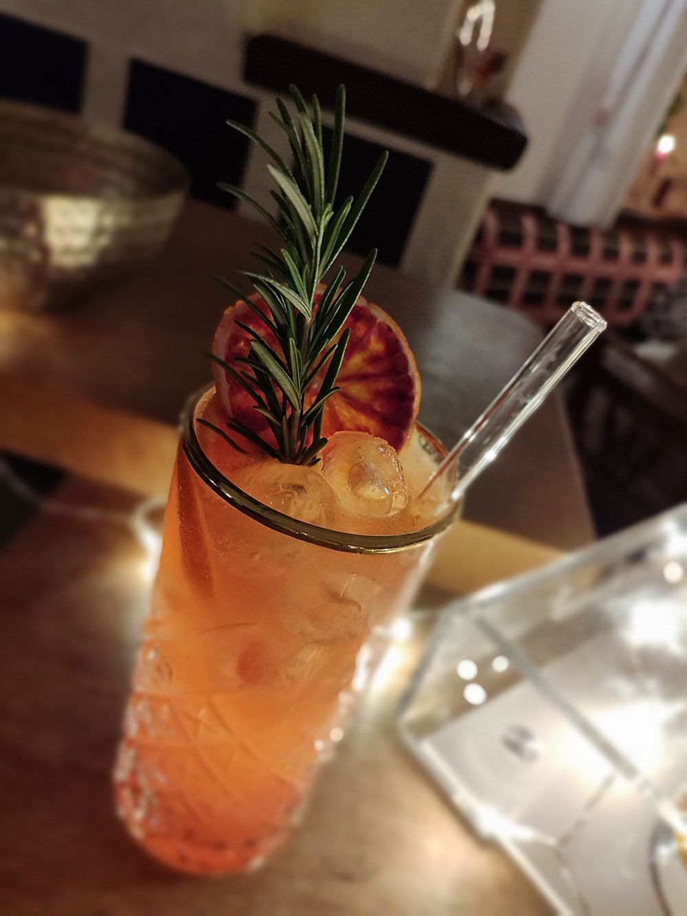Orangencocktail in einem hohen Glas, garniert mit Rosmarin und Blutorangenscheibe, auf einer Holzoberfläche.