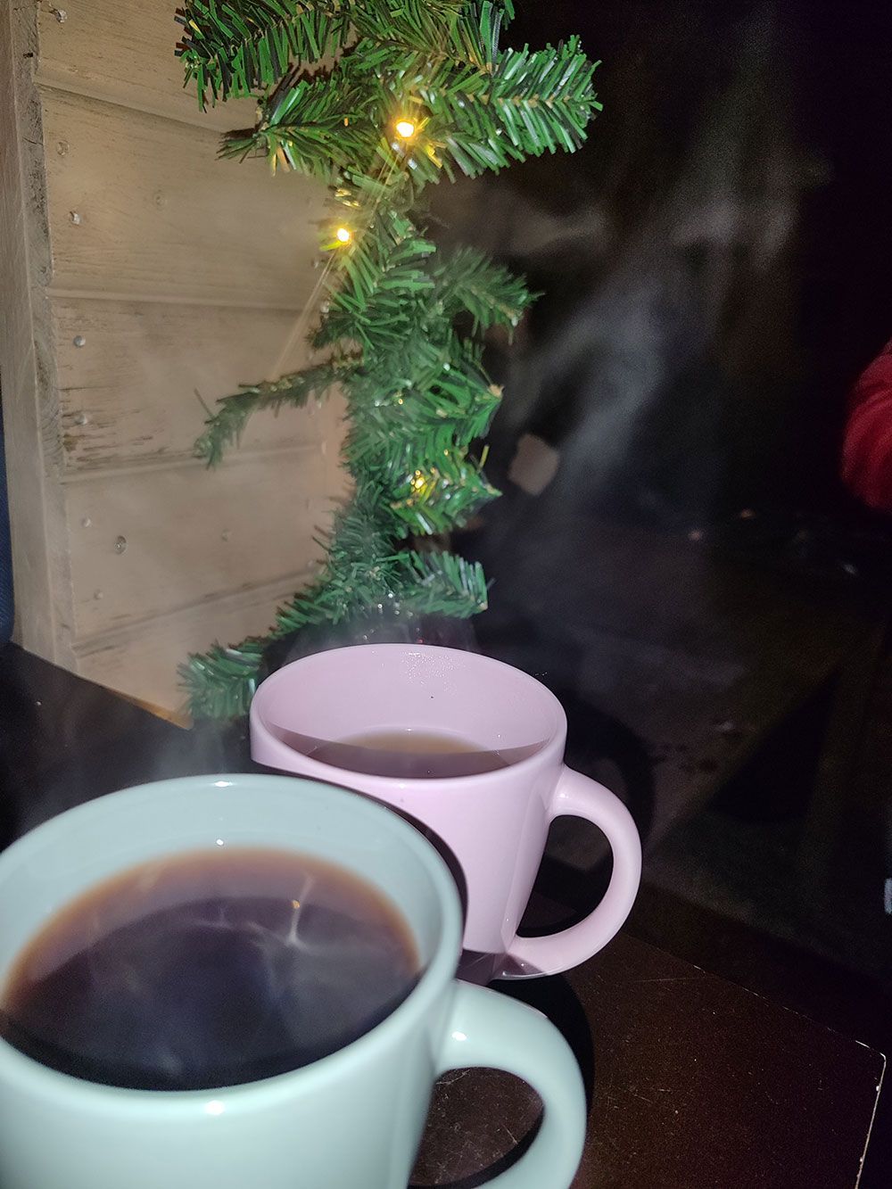 Zwei Tassen dampfender Kaffee neben einem kleinen beleuchteten Weihnachtsbaum auf einer dunklen Tischplatte.
