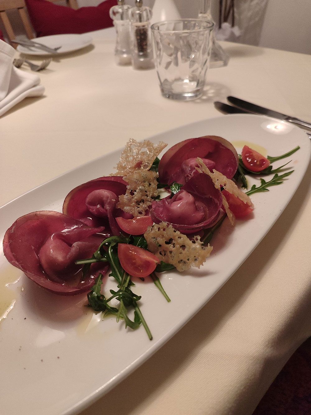 Rote-Bete-Ravioli-Vorspeise auf weißem Teller, Rucola, Tomaten und Käsechips auf weißem Tischtuch.