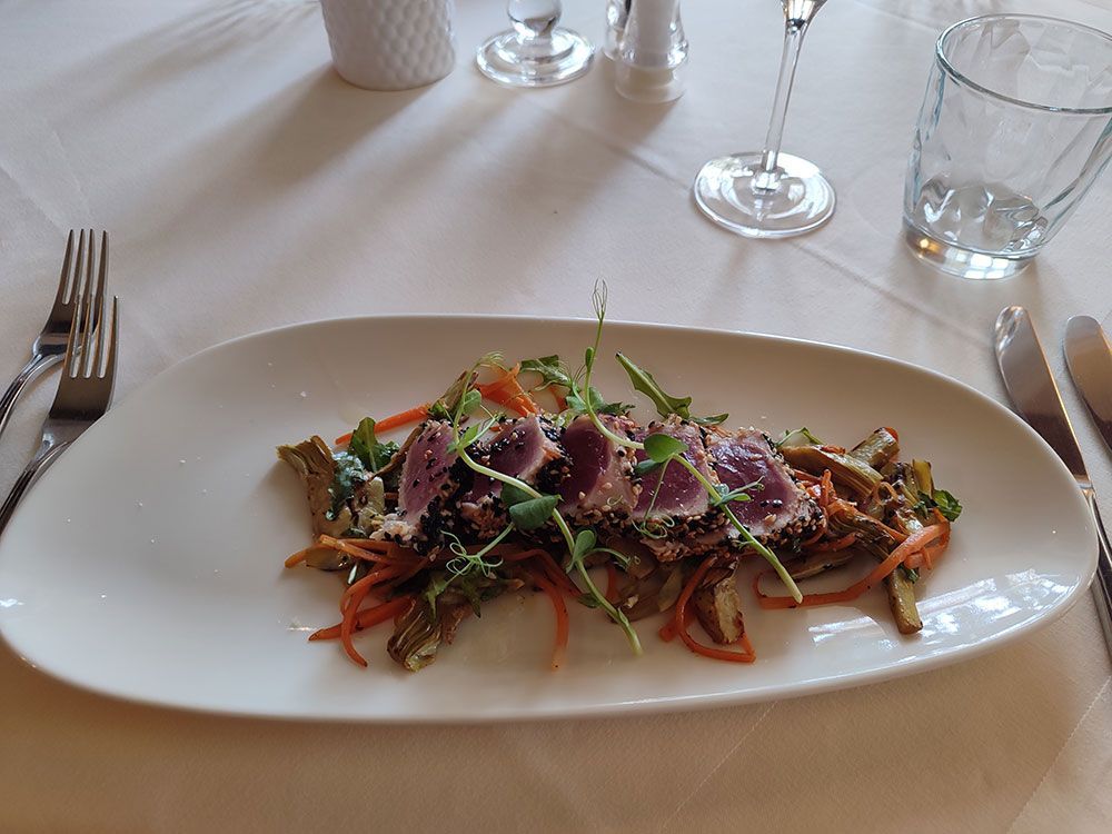 Gebratener Thunfisch mit Sesamkörnern auf einem Gemüsebett, serviert auf einem weißen Teller an einem gedeckten Tisch.