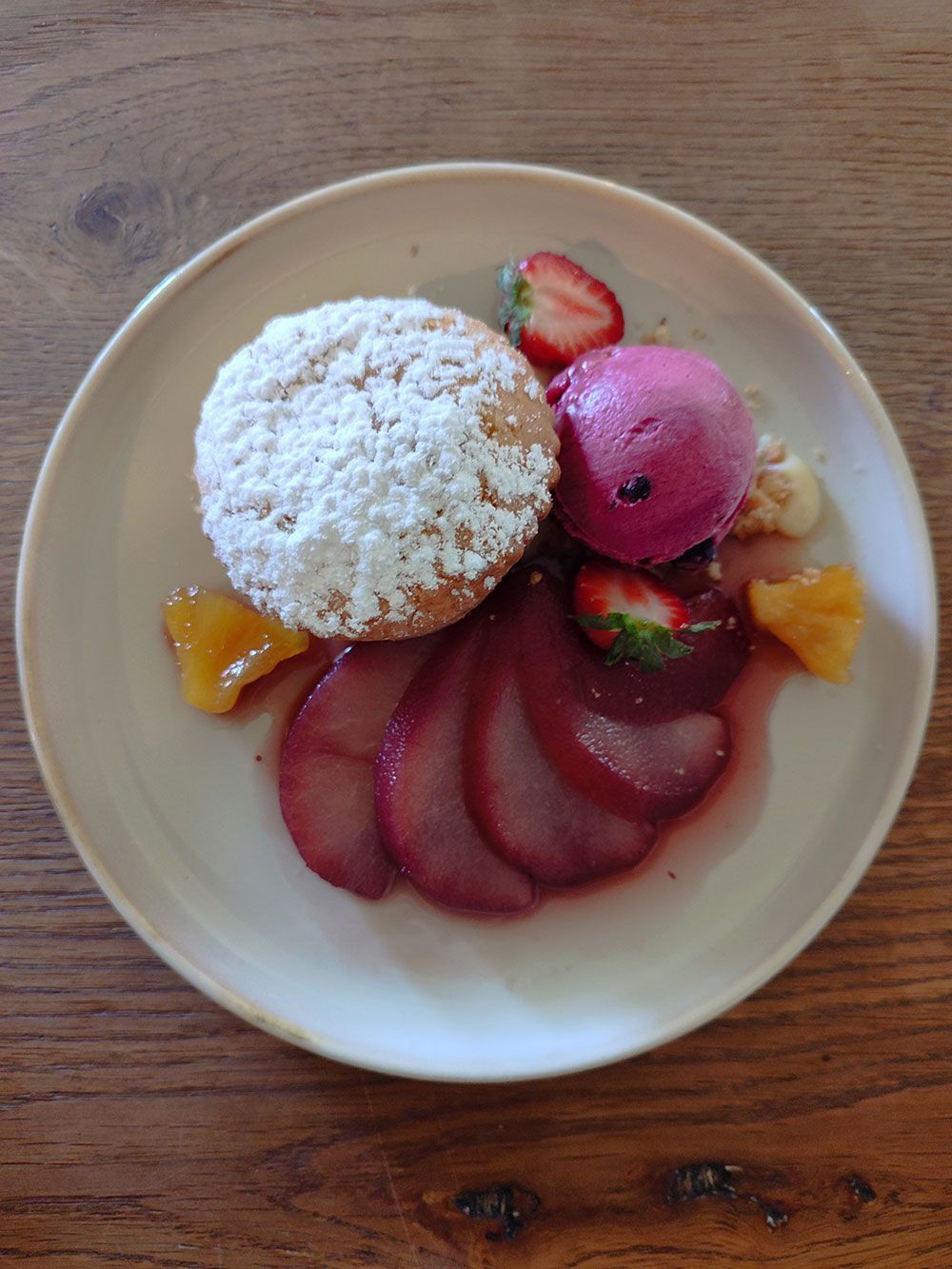 Dessertteller mit einem mit Puderzucker bestäubten Muffin, pochierten Birnenscheiben, rosa Eis, Erdbeeren und Orangenscheiben.