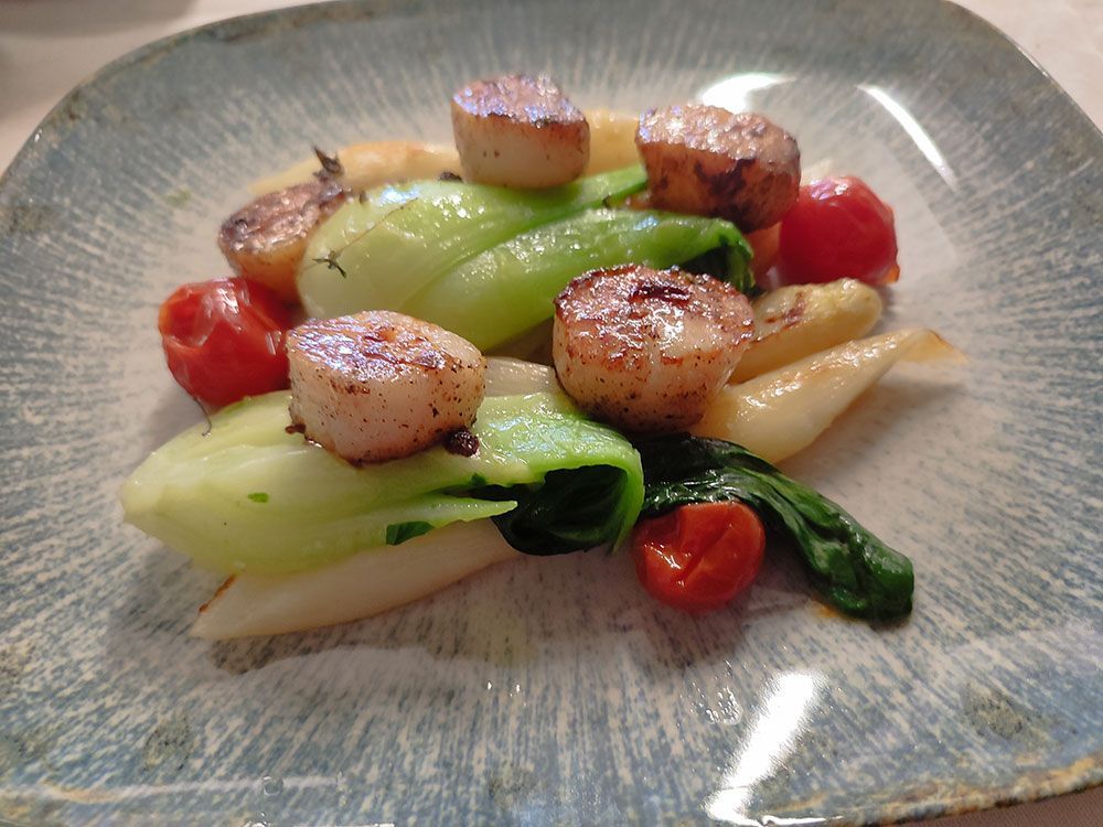 Gebratene Jakobsmuscheln mit Pak Choi, Tomaten und weißem Spargel auf einem blaugrauen Teller.