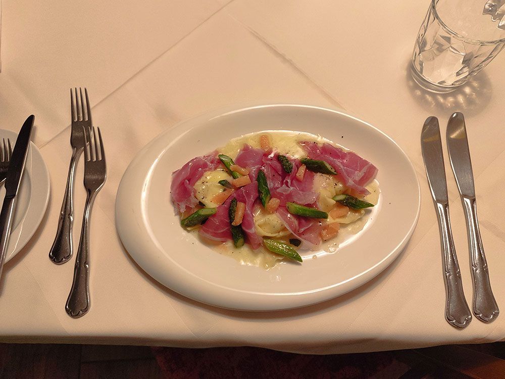Teller mit Ravioli mit Spargel und Prosciutto auf einem mit Besteck und einem Wasserglas gedeckten Tisch.