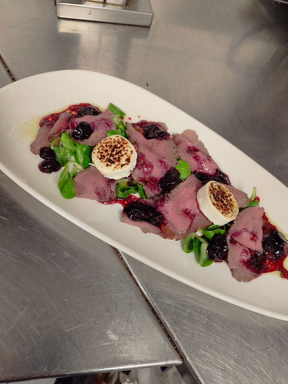 Fleischscheiben mit Blaubeerkompott, Gemüse und geröstetem Ziegenkäse auf einer weißen Platte.