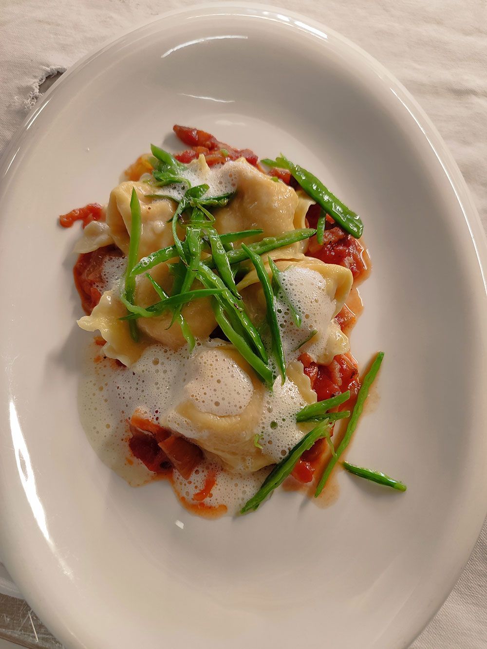 Nudelgericht mit grünen Bohnen, Tomaten und Schaum, serviert auf einem weißen ovalen Teller.