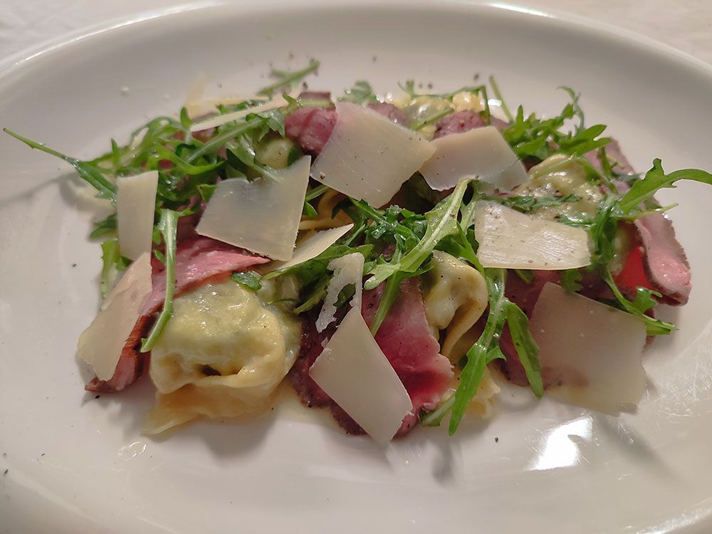 Nudelsalat mit Rucola, gehobeltem Parmesan und Aufschnitt auf einem weißen Teller.