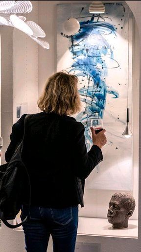 Eine Frau betrachtet in einem Museum ein Gemälde und hält dabei ein Glas Wein in der Hand.