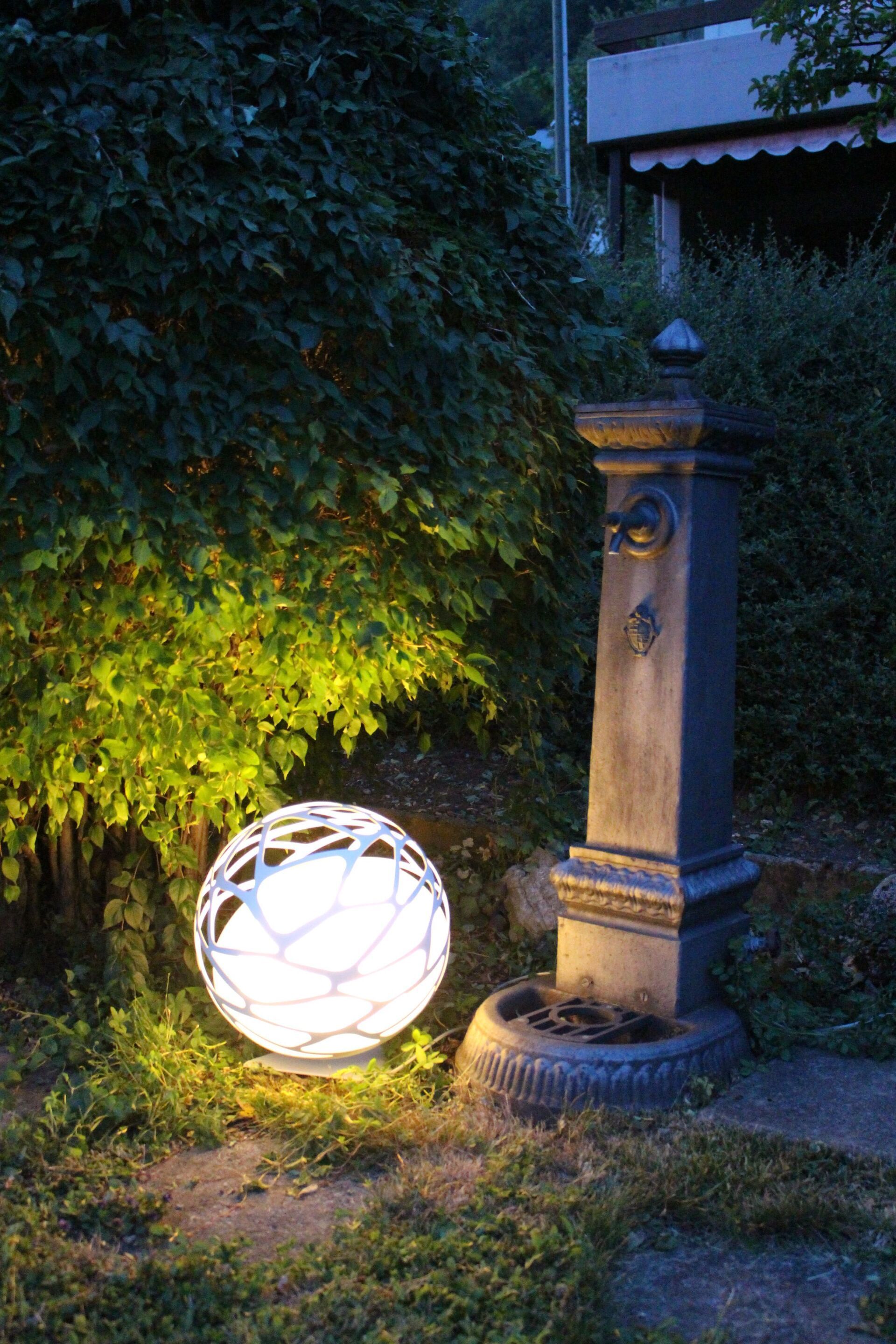 In einem Garten ist nachts ein Brunnen beleuchtet, im Vordergrund steht eine Kugel.