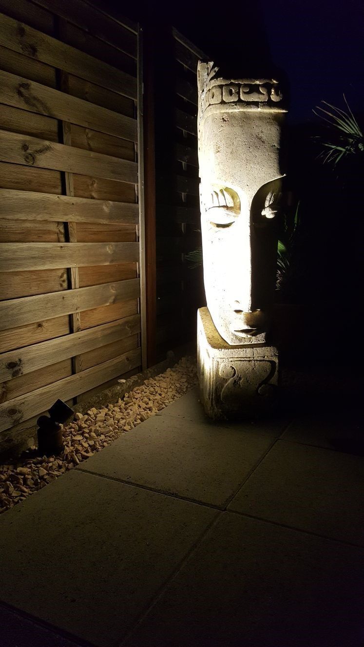 Vor einem Holzzaun wird nachts eine Gesichtsstatue beleuchtet.