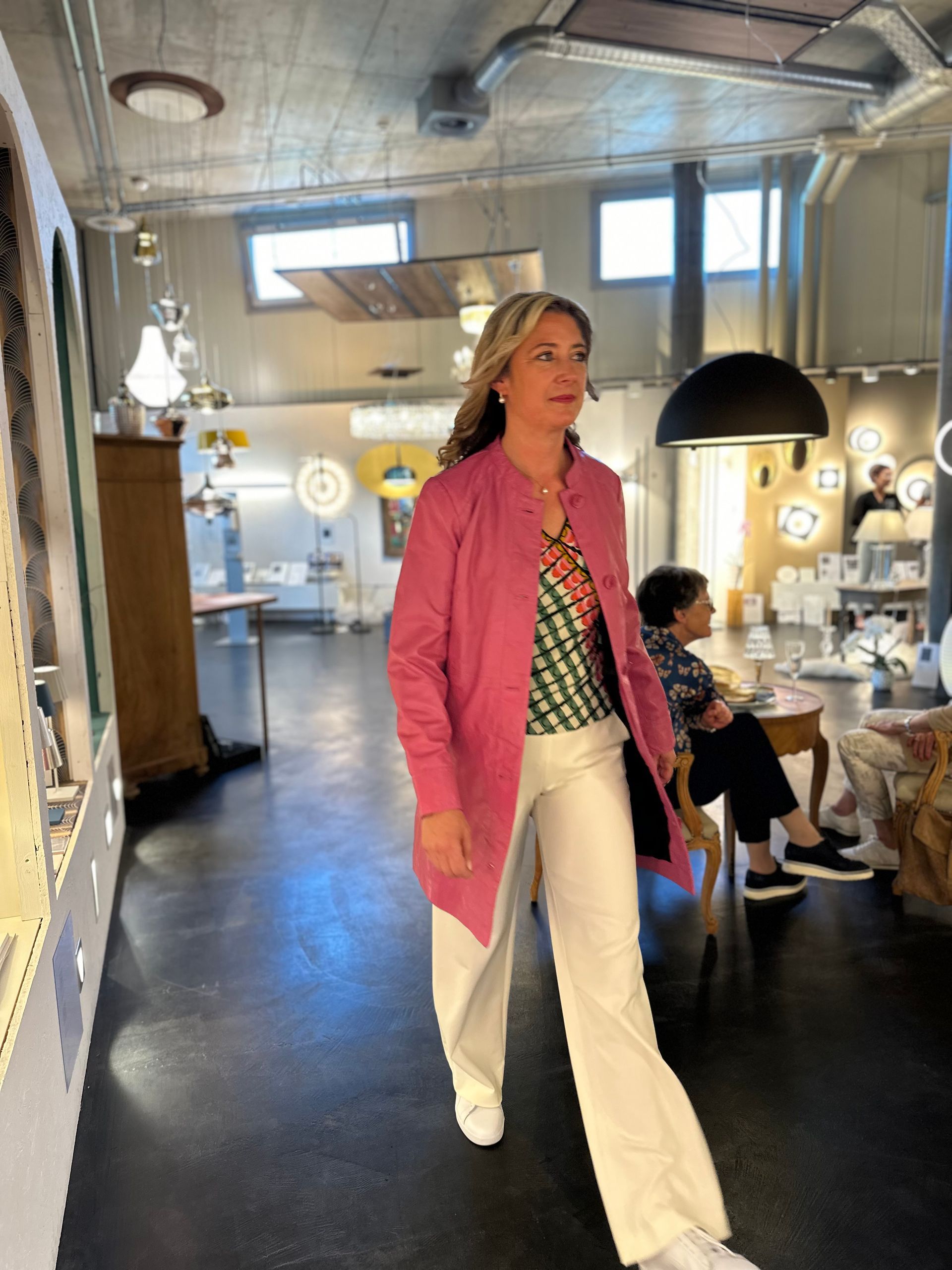 Eine Frau in einer rosa Jacke und weißen Hosen geht in einen Raum.