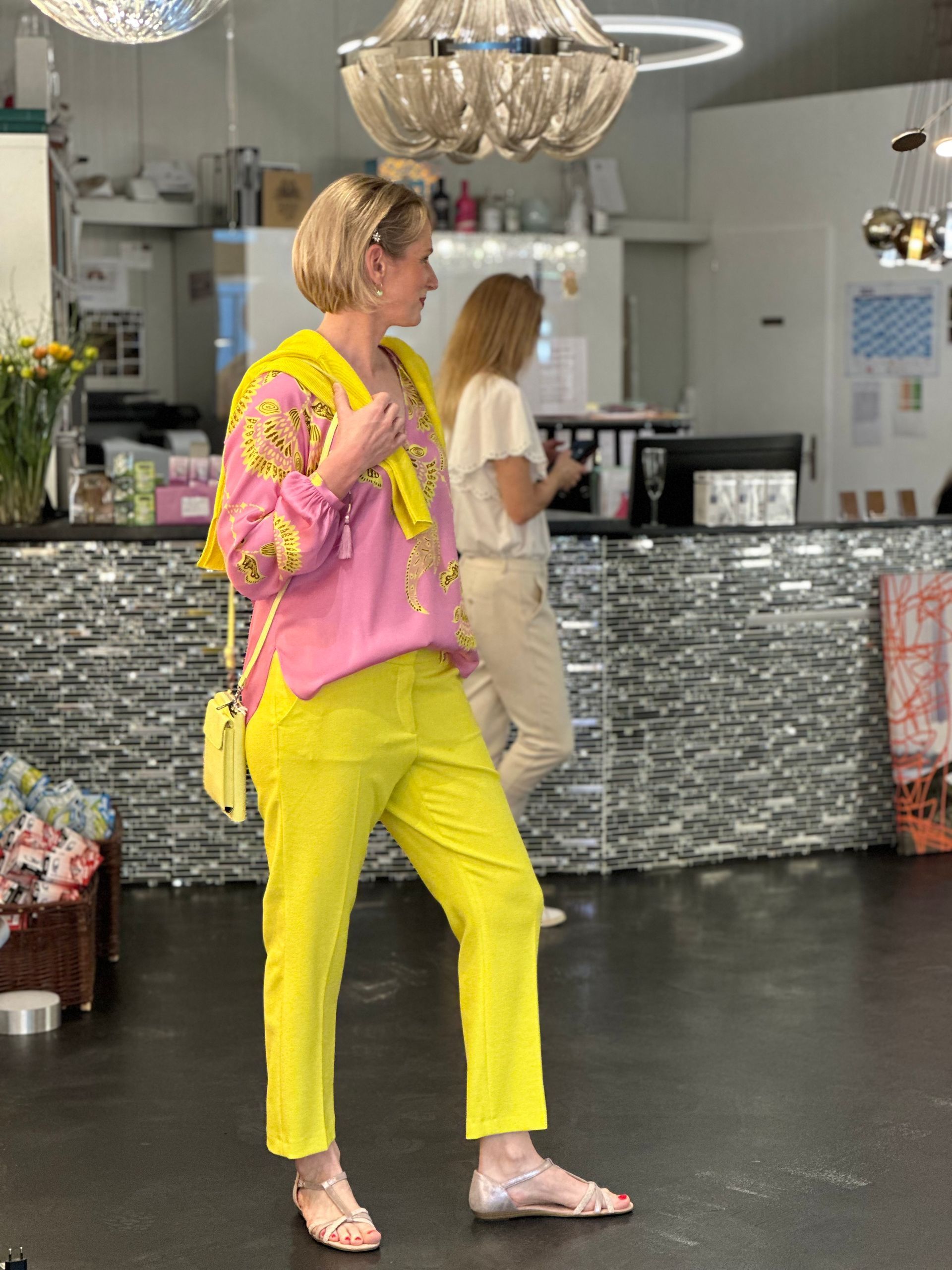 Eine Frau in einem rosa Hemd und gelben Hosen geht in ein Geschäft.