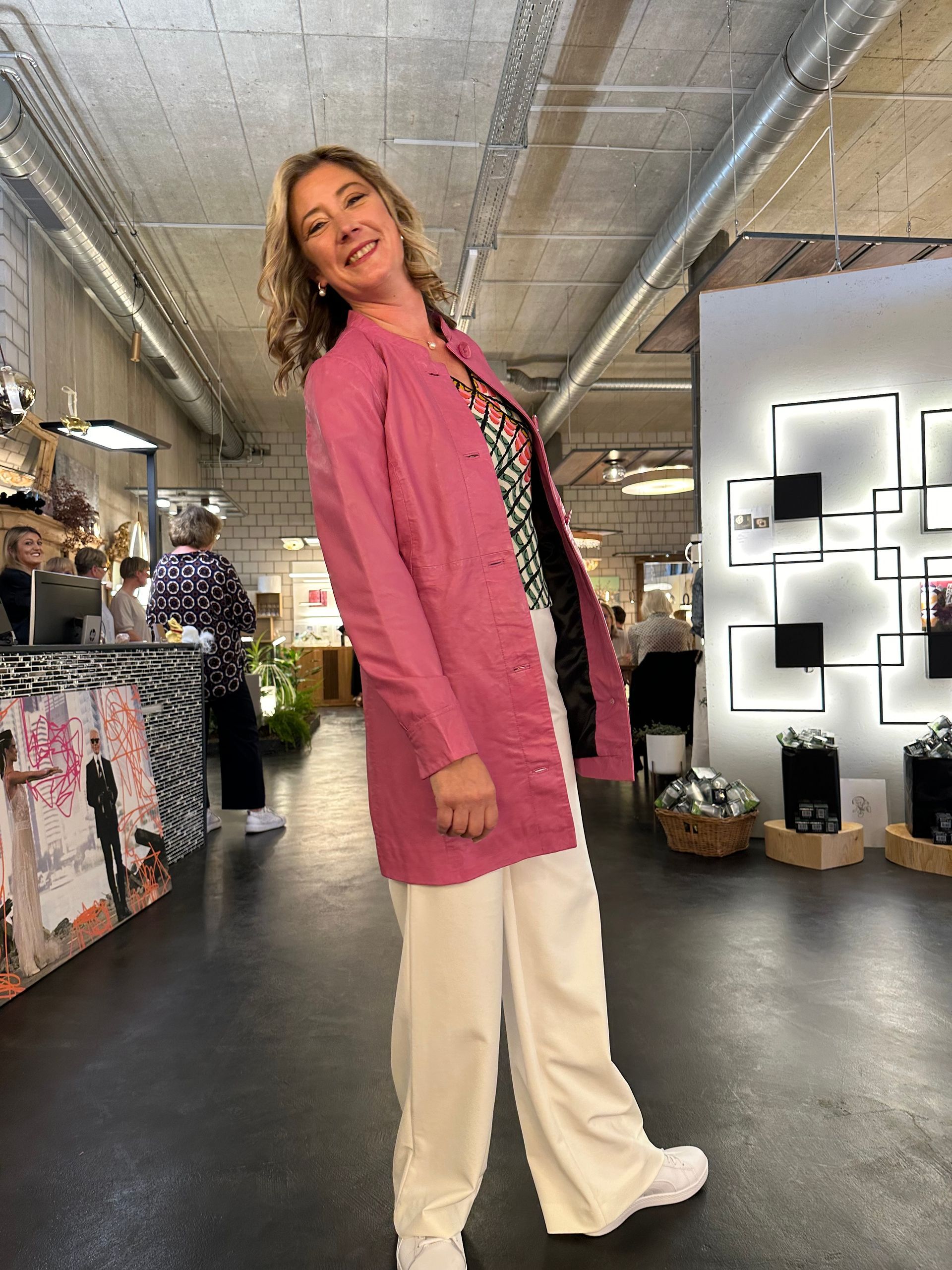 Eine Frau in einer rosa Jacke und weißen Hosen steht in einem Raum.