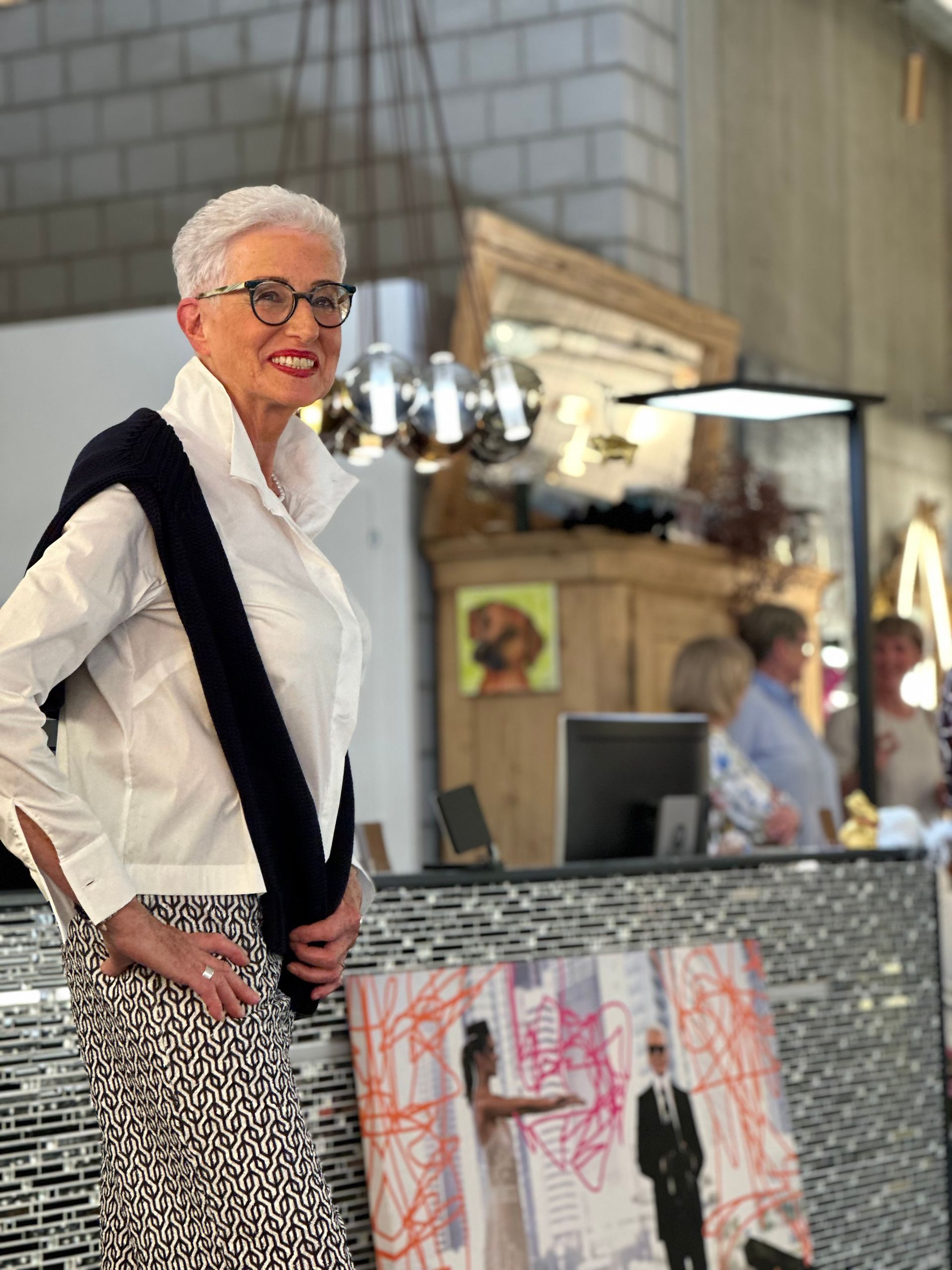 Eine Frau mit Brille und weißem Hemd steht vor einem Gemälde.