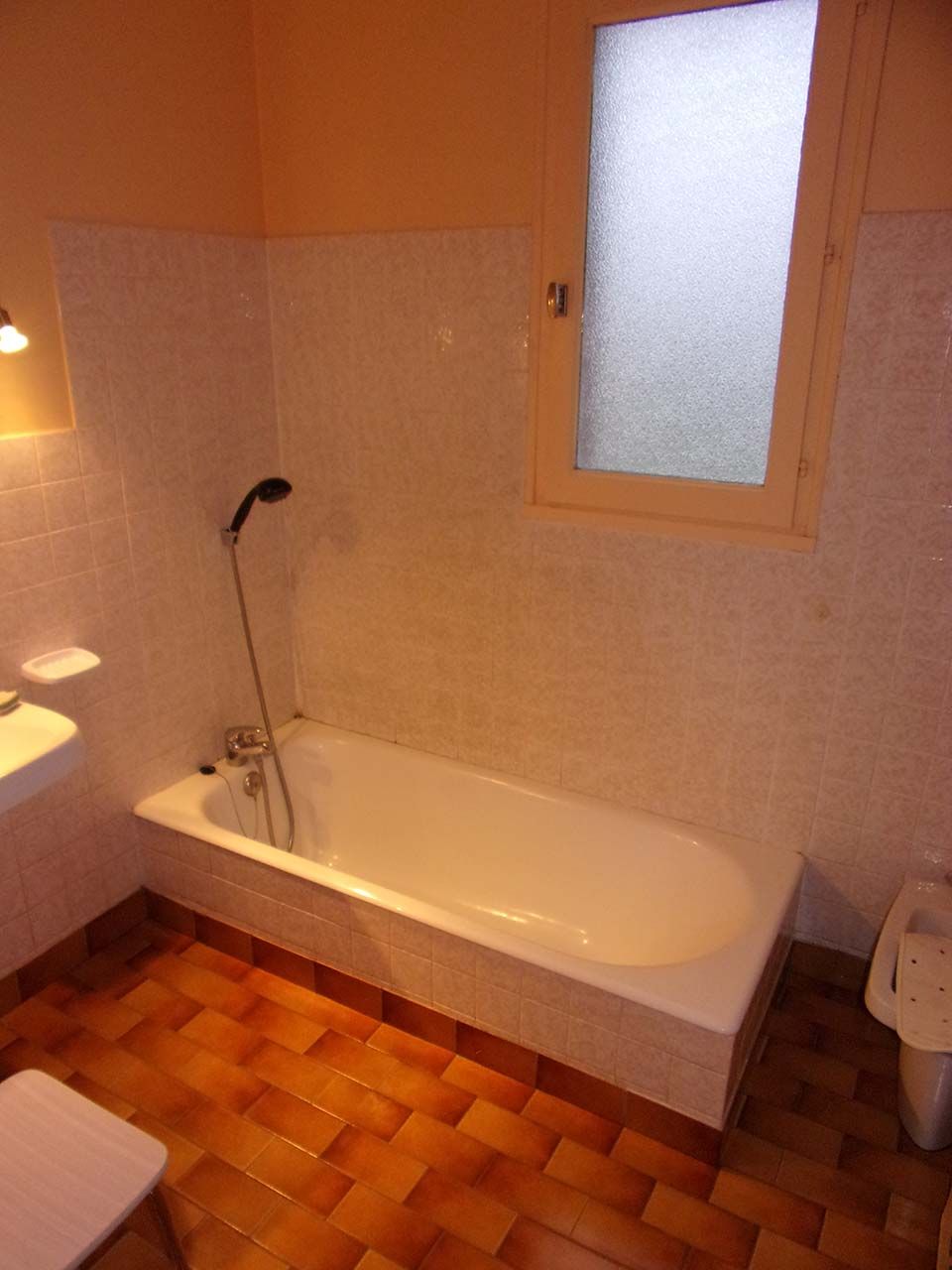 Une salle de bains avec une baignoire blanche, une douchette, des murs carrelés clairs et un sol carrelé marron.