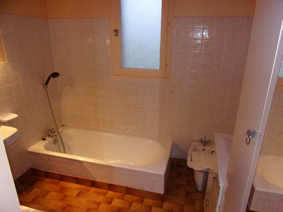 Une salle de bains avec baignoire blanche, douchette, bidet et murs carrelés clairs sur un sol carrelé marron.