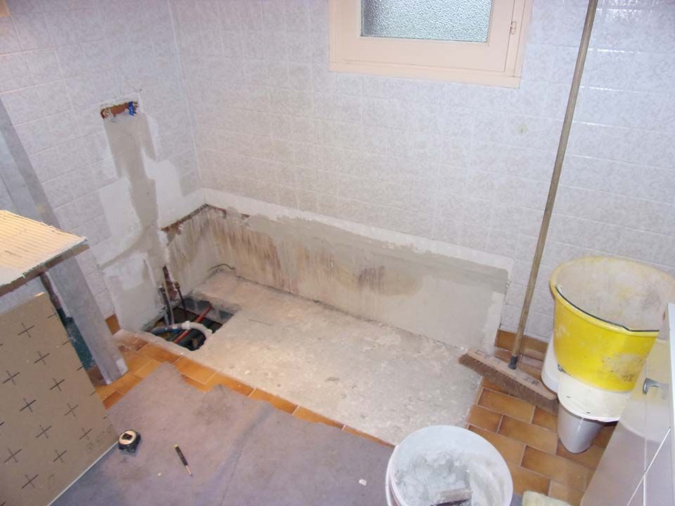 Une salle de bains en cours de rénovation avec des tuyaux de plomberie apparents, une section de sol en béton et des seaux de chantier.
