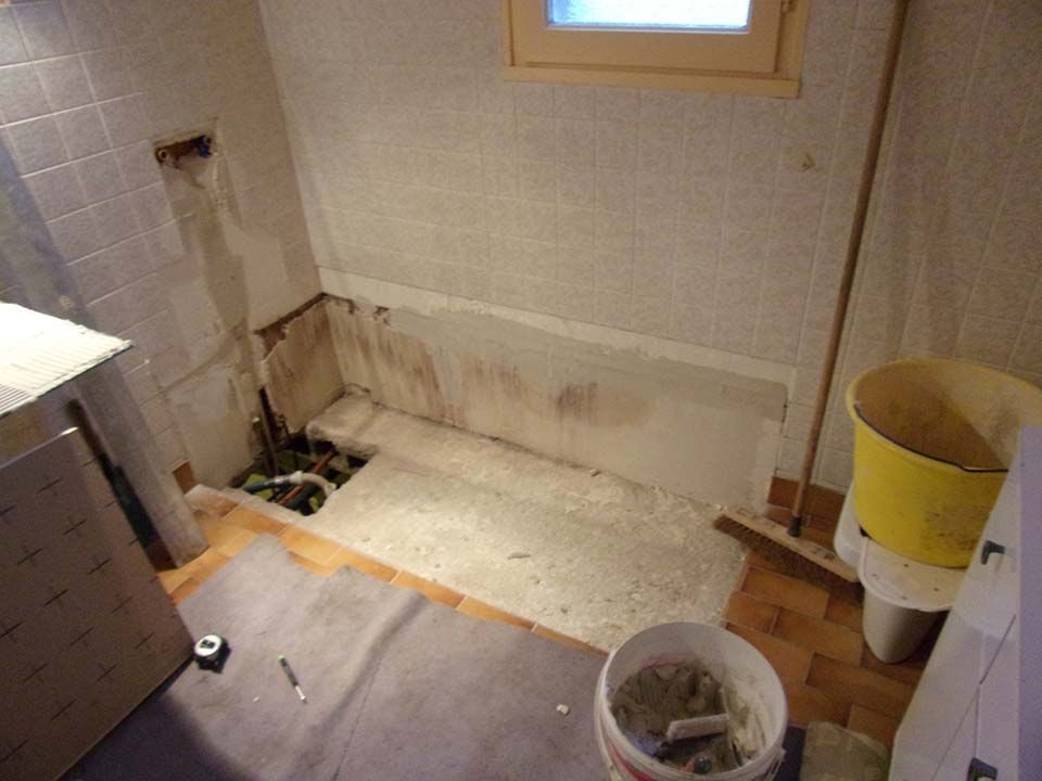 Rénovation en cours d'une salle de bain intérieure : la baignoire a été retirée, laissant apparaître la plomberie, les murs carrelés et les outils de chantier.
