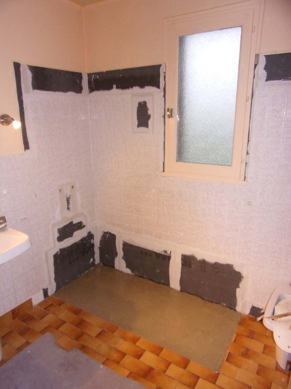 Une salle de bains en cours de rénovation avec des murs blancs partiellement carrelés, du mortier gris apparent et un parquet en bois.