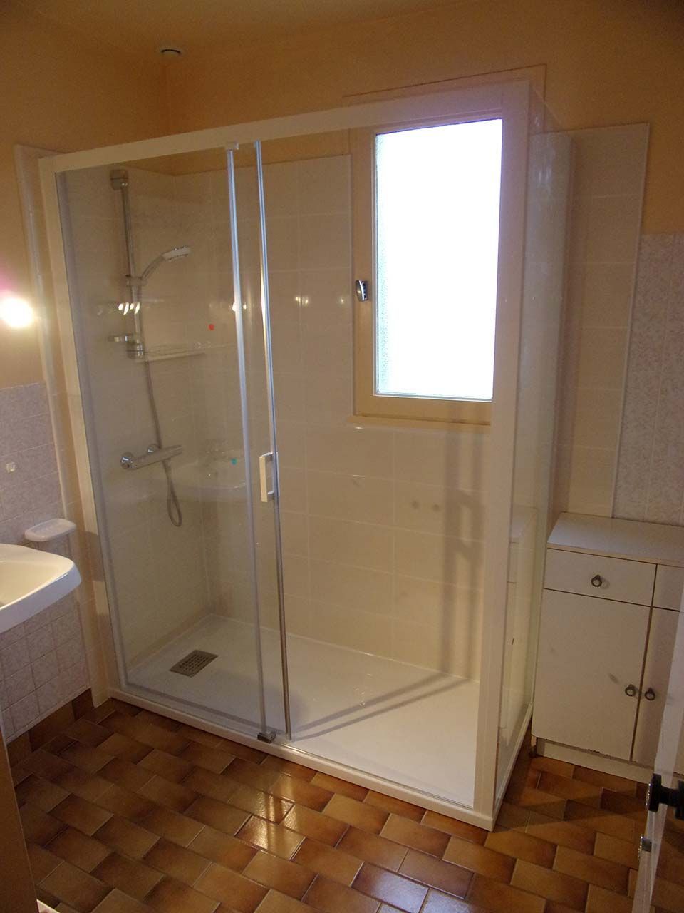 Une cabine de douche avec une porte en verre et un cadre blanc, située dans une salle de bains au sol carrelé et dotée d'une petite fenêtre.