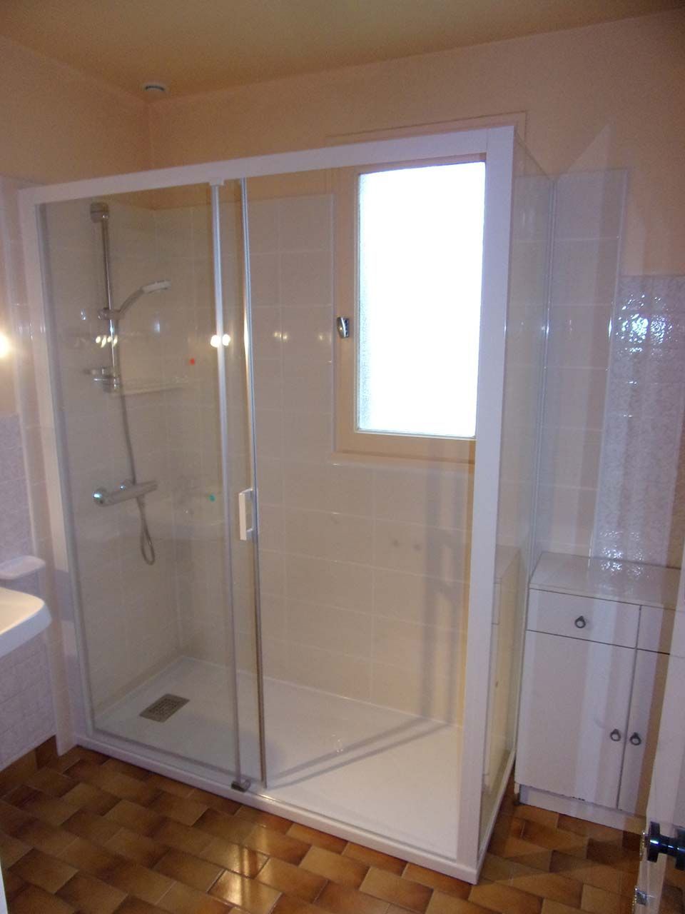 Une cabine de douche en verre à cadre blanc dans une salle de bains avec fenêtre, meuble-lavabo blanc et sol carrelé brun clair.
