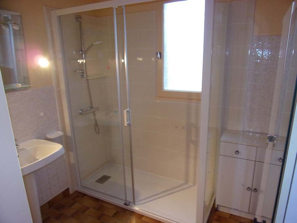 Une salle de bains lumineuse avec un lavabo sur pied, une cabine de douche vitrée et une petite armoire de rangement blanche.