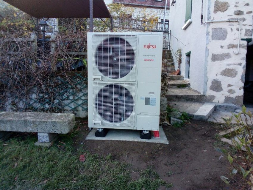 Une unité de pompe à chaleur extérieure Fujitsu, montée sur une base en béton dans un jardin jouxtant une maison en pierre.