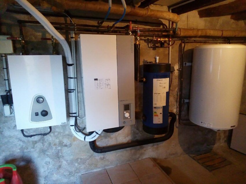 Une salle des machines montrant une chaudière blanche, un boîtier de commande gris, un vase d'expansion bleu et un grand réservoir d'eau blanc.