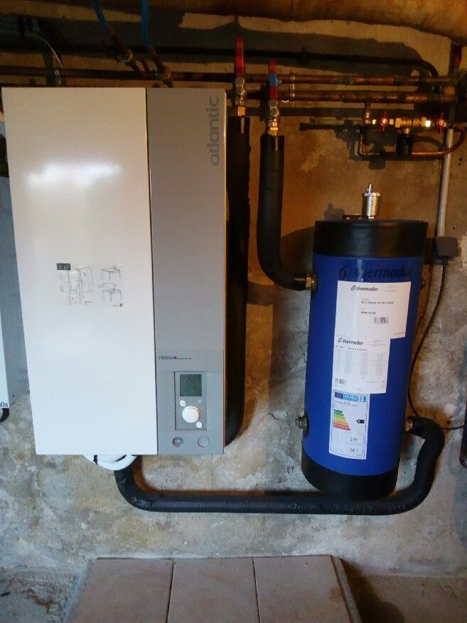 Une pompe à chaleur Atlantic blanche et grise est placée à côté d'un chauffe-eau cylindrique bleu, fixé sur un mur en pierre brute.