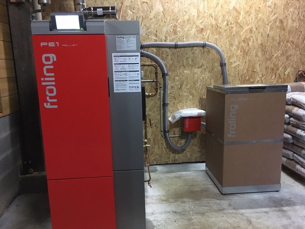 Une chaudière à granulés Froling rouge reliée par des tuyaux à une grande trémie à combustible brune et cubique, dans une pièce aux murs lambrissés.