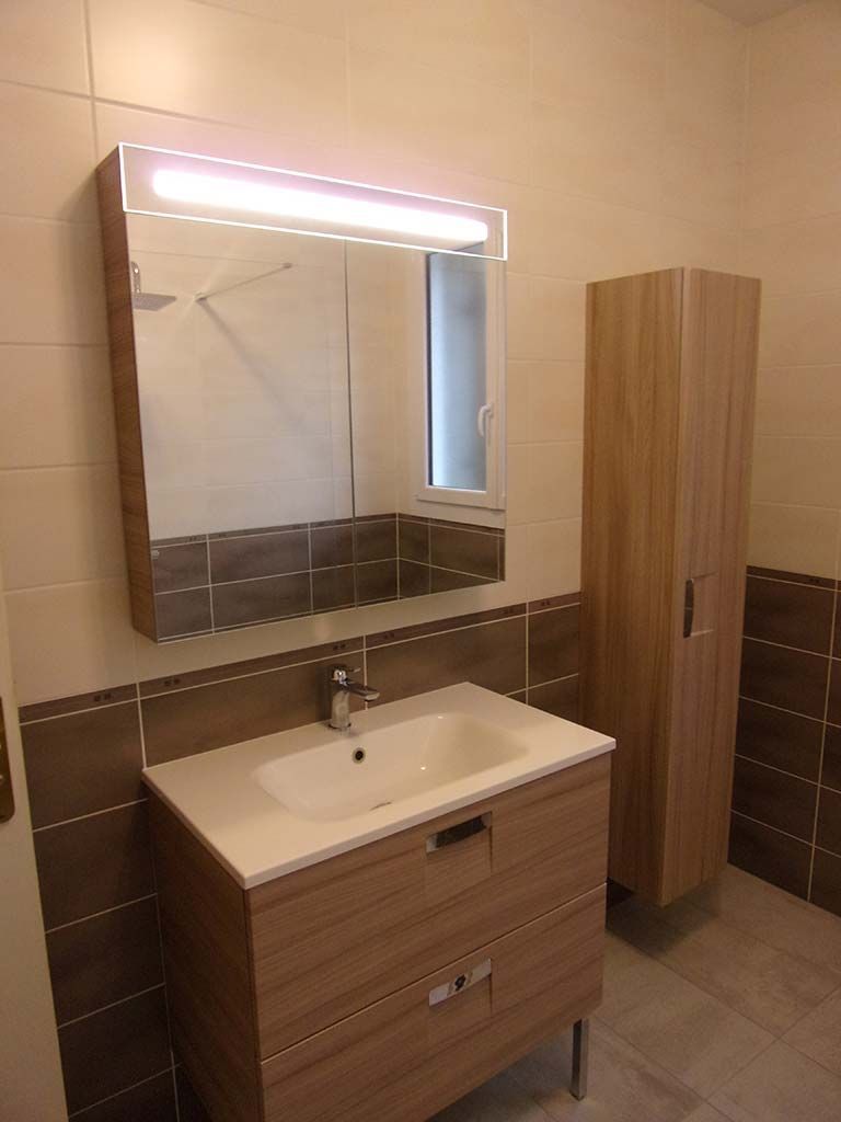 Un meuble-lavabo moderne avec un miroir éclairé au-dessus d'un lavabo blanc et une haute armoire couleur bois sur la droite.