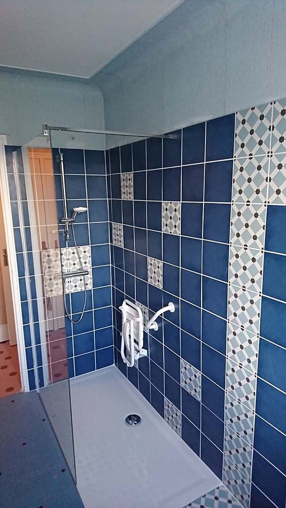 Une douche à l'italienne avec des carreaux carrés bleus, des éléments décoratifs, un siège de douche rabattable blanc et une barre d'appui.