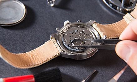 Uhr wird repariert, mit einer Pinzette wird am inneren Mechanismus gearbeitet. Werkzeuge, Uhrenarmband und Teile auf einer dunklen Oberfläche.