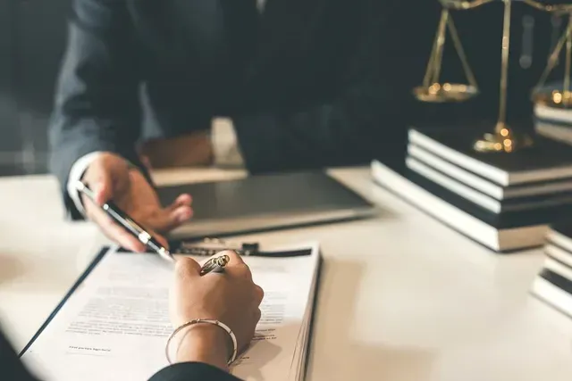 Una persona firma un documento mientras otra persona lo señala, con una regla legal y libros sobre un escritorio cercano.