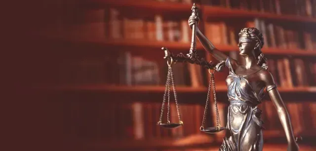 Una estatua de bronce de la Dama de la Justicia sosteniendo una balanza, situada frente a un fondo borroso de estanterías.