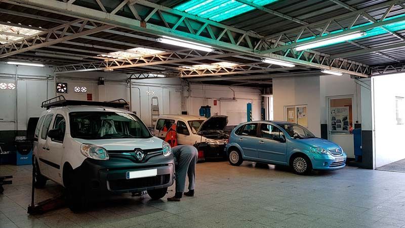 Coches en un taller mecánico. Mecánico trabajando en una furgoneta blanca. Coche azul y otro vehículo visibles.