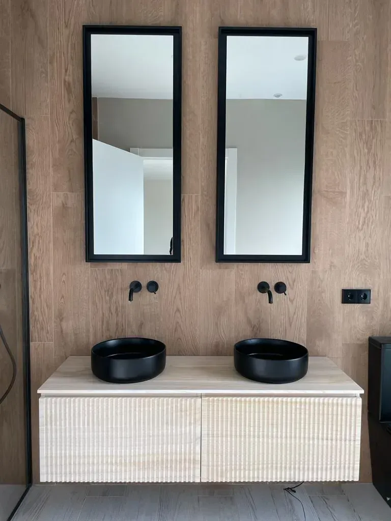 Baño con mueble de madera, dos lavabos negros y espejos, sobre pared efecto madera.