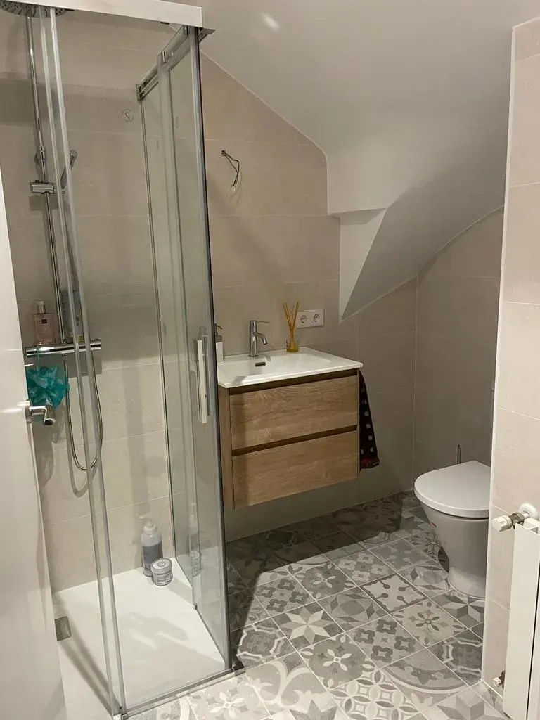 Pequeño baño con ducha, tocador e inodoro; azulejos grises y beige.