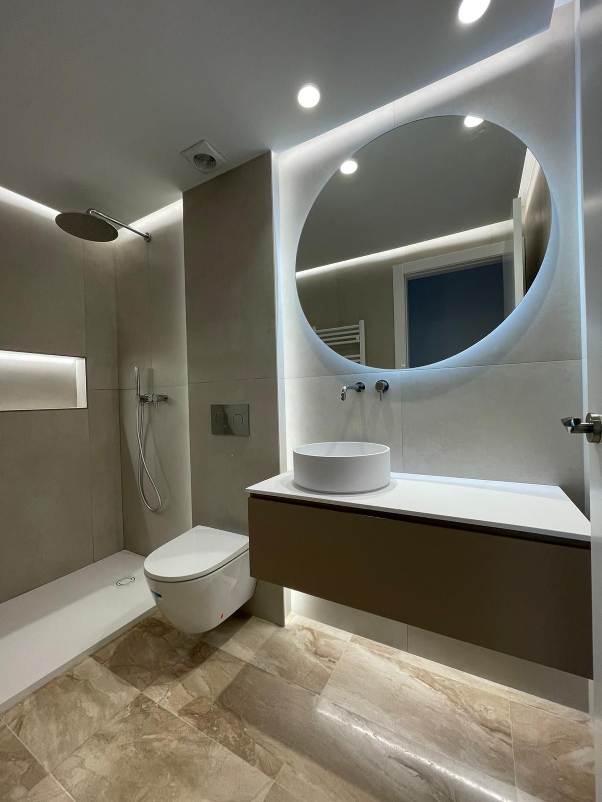 Baño con espejo ovalado retroiluminado, azulejos en espiga gris, lavabo blanco y tocador de madera.