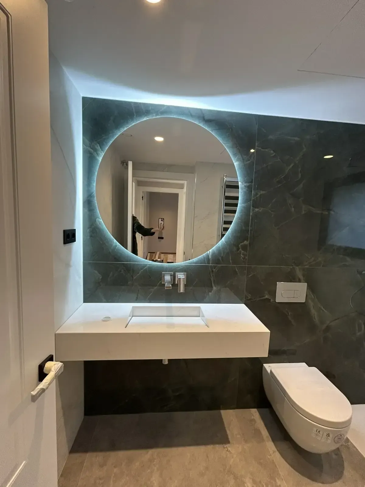 Baño moderno con espejo redondo iluminado, lavabo flotante, inodoro y pared con apariencia de mármol.
