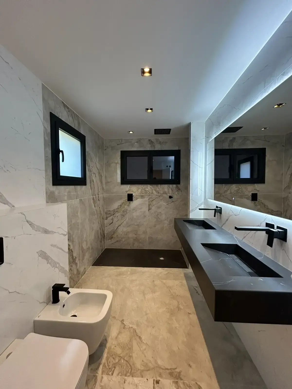 Baño moderno con paredes y suelo de mármol, accesorios negros y lavabos dobles.