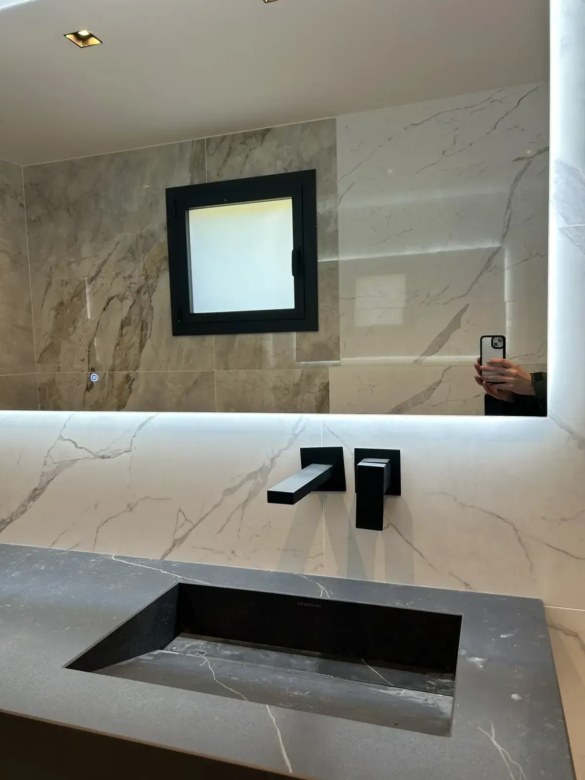 Baño con lavabo cuadrado negro, grifo y espejo. Paredes de mármol blanco con un espejo cuadrado más pequeño insertado.