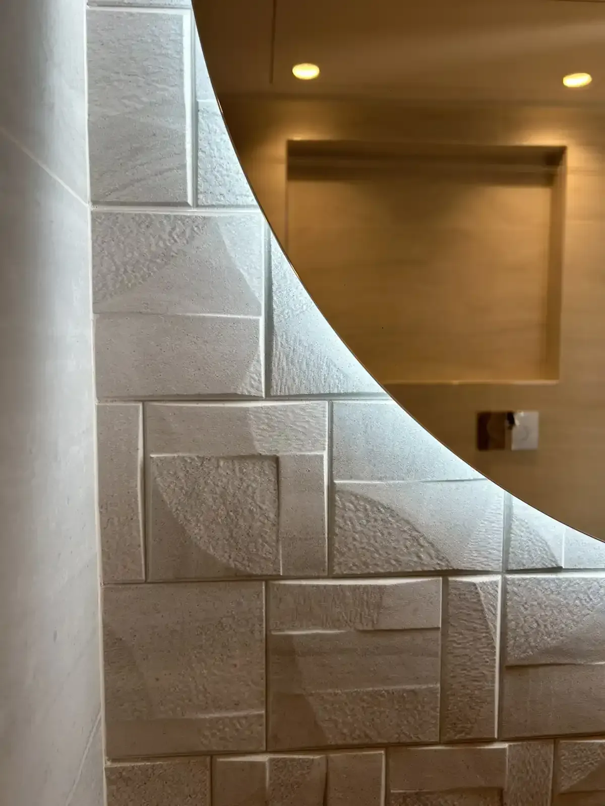 Pared blanca texturizada con patrones geométricos, espejo curvo parcialmente visible, entorno en tonos neutros.
