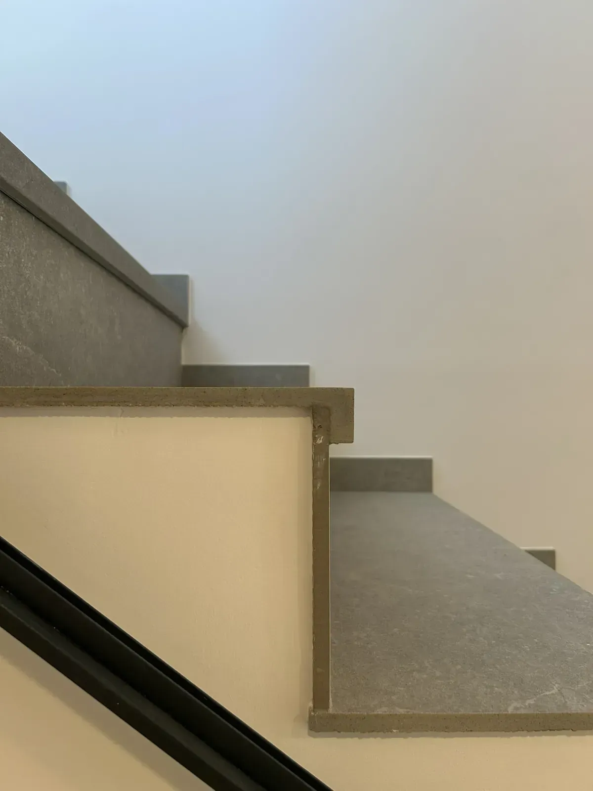 Peldaños de escalera de piedra gris contra paredes blancas; pasamanos negro visible.