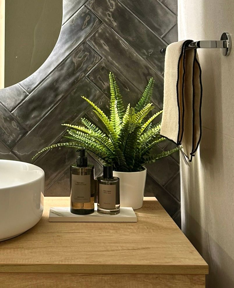 Baño con espejo ovalado retroiluminado, azulejos en espiga gris, lavabo blanco y tocador de madera.