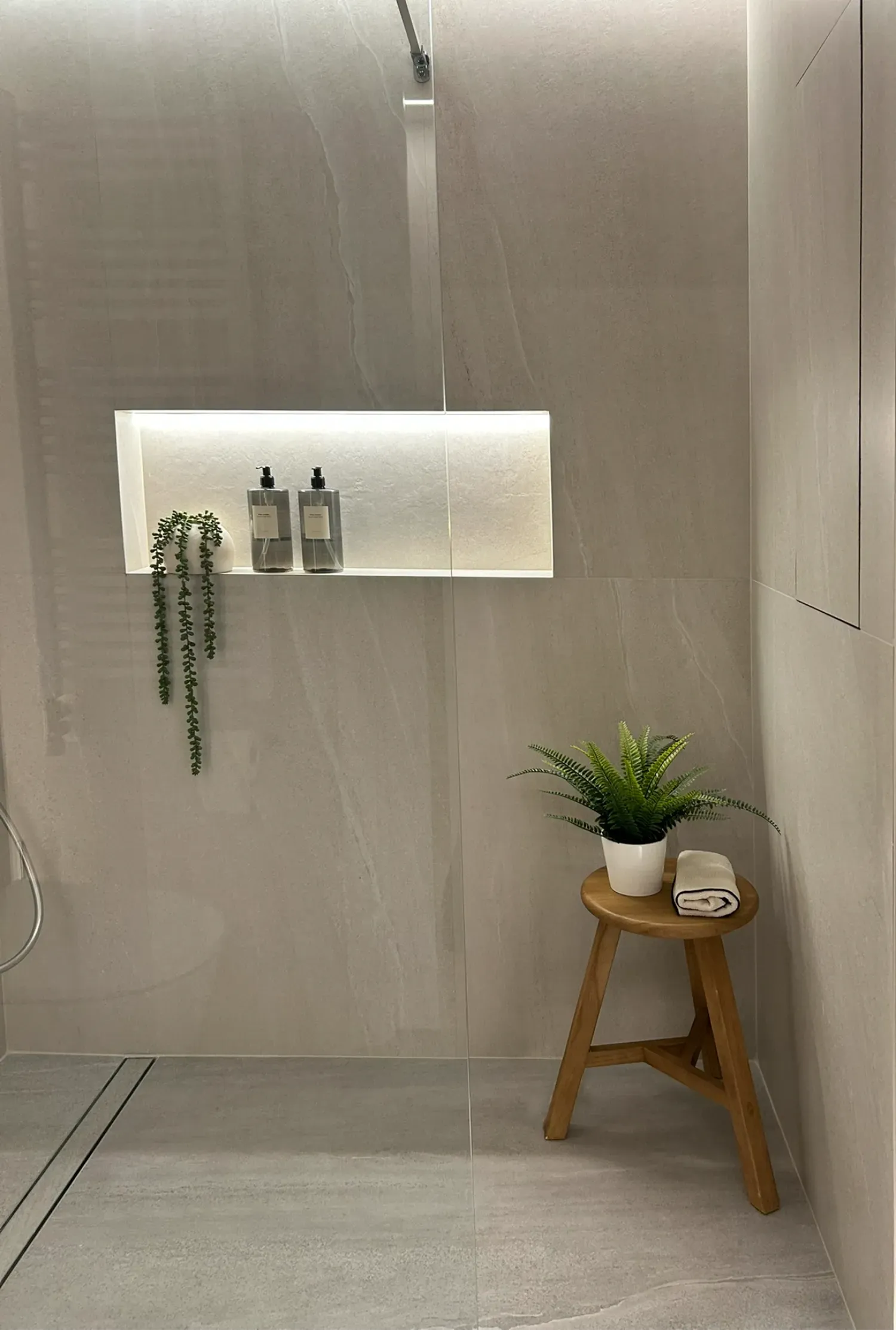Ducha con paredes de azulejos gris claro. Estante con nicho iluminado y artículos de aseo. Taburete de madera con planta.