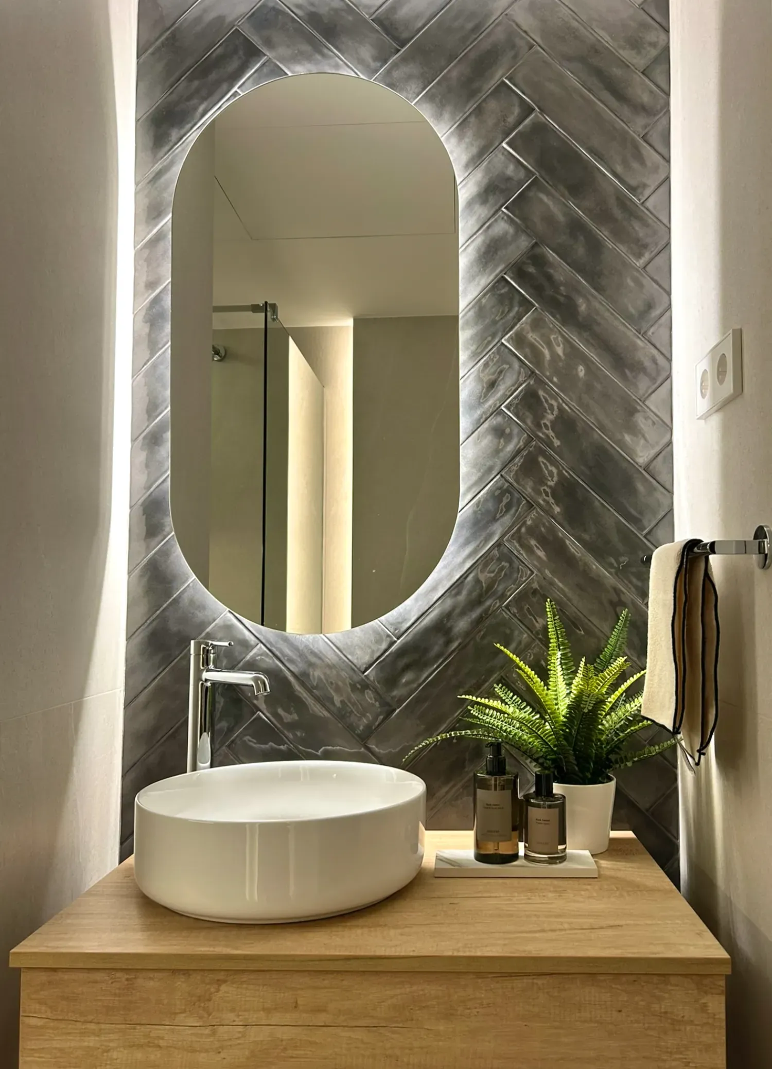 Baño con espejo ovalado retroiluminado, azulejos en espiga gris, lavabo blanco y tocador de madera.