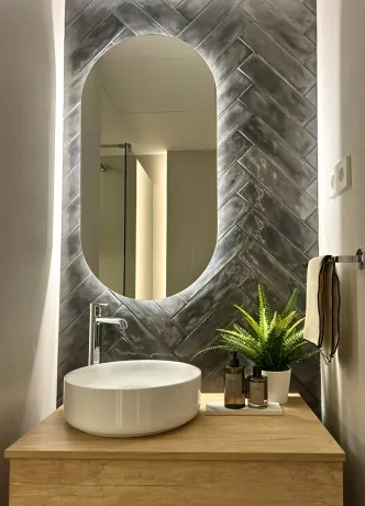 Baño con espejo ovalado retroiluminado, azulejos en espiga gris, lavabo blanco y tocador de madera.