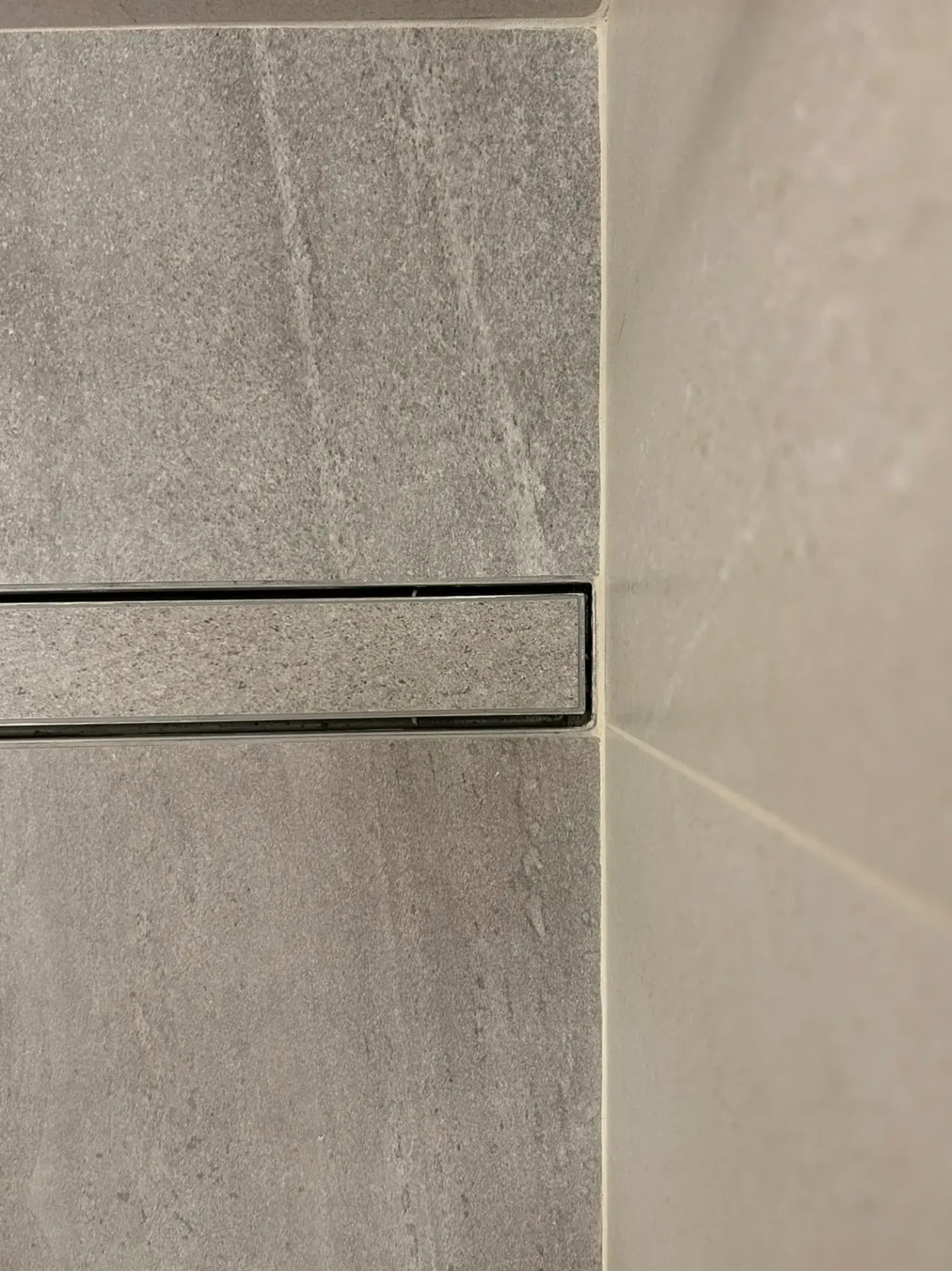 Un desagüe de ducha lineal de color negro entre paredes de azulejos grises y blanquecinos.