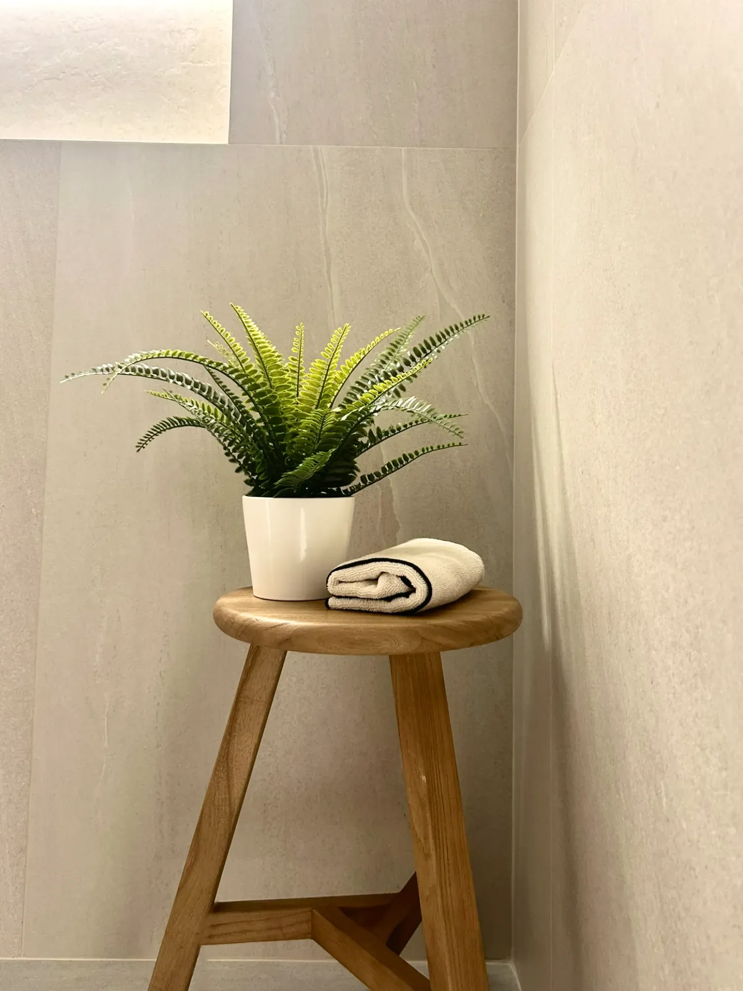 Una planta verde en maceta y una toalla enrollada descansan sobre un taburete de madera en un baño de tonos claros.
