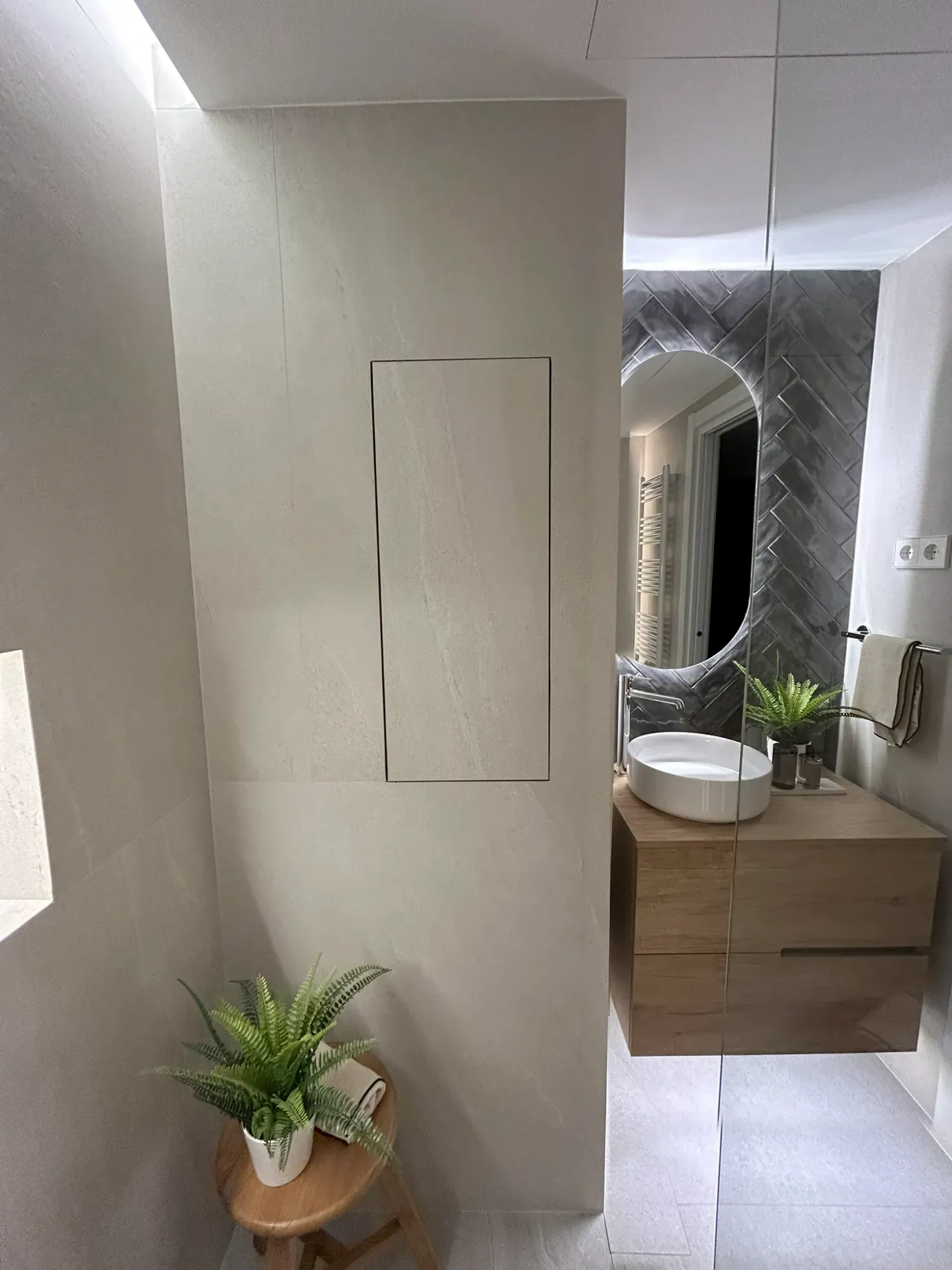 Baño moderno con tocador de madera clara, espejo redondo y un pequeño taburete con una planta.